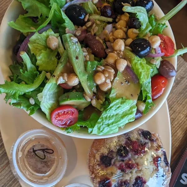 A Green Beans Coffee salad with avocado, chickpeas, olives, and tomatoes, next to a berry muffin.