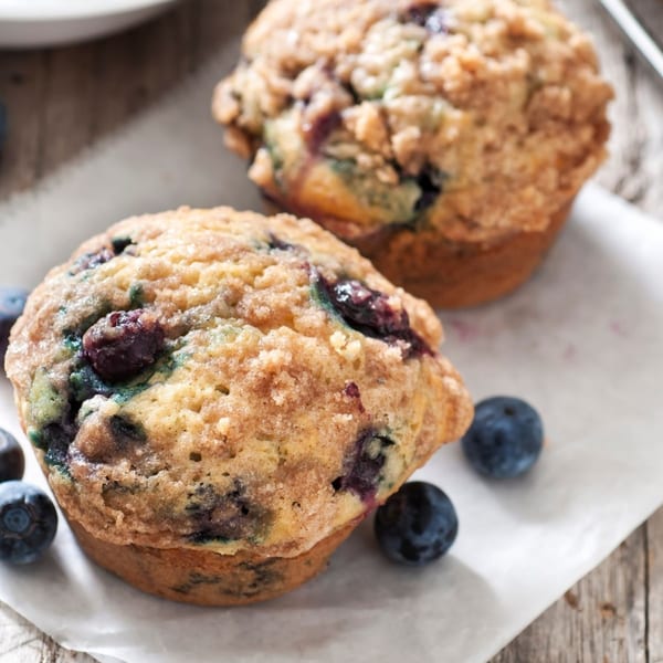 Two blueberry muffins with crumb topping and fresh blueberries on white parchment paper.