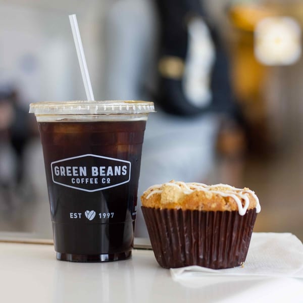 Green Beans Coffee iced coffee in a branded cup next to a muffin with white icing.