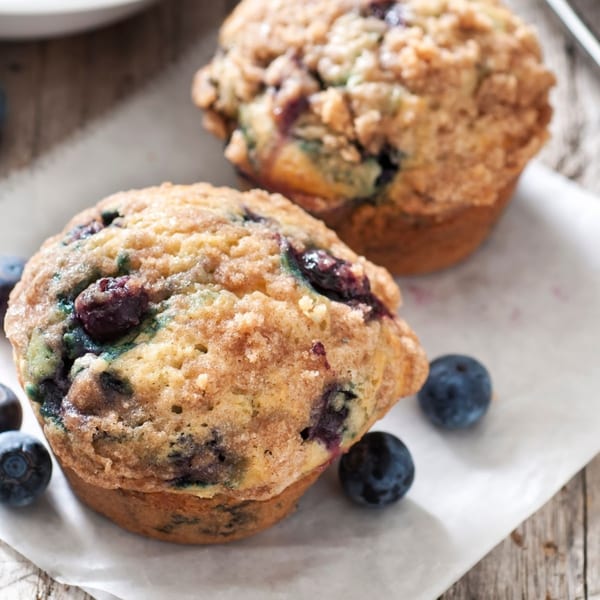 Two blueberry muffins with crumb topping and fresh blueberries on parchment paper.