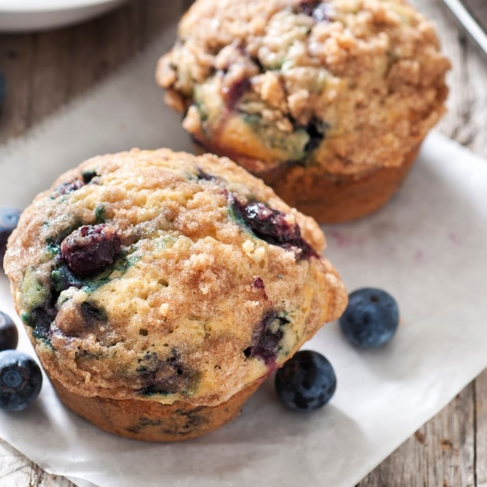 Two blueberry muffins with crumb topping and fresh blueberries on parchment paper.