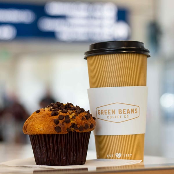 A Green Beans Coffee cup and a chocolate chip muffin sit on a white napkin.