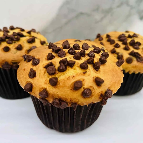 Three golden-brown chocolate chip muffins in dark brown wrappers, with a blurred background.