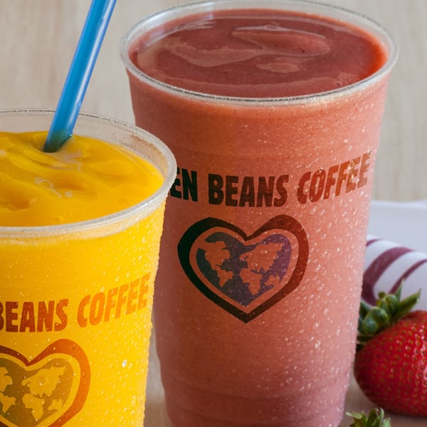 Two Green Beans Coffee smoothies, one yellow and one red, with fresh strawberries nearby.